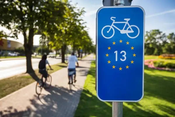Znak EuroVelo 13 na ścieżce rowerowej. Dwie osoby jadą na rowerach wzdłuż alei drzew.
