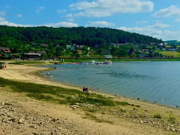 Słoneczna plaża nad Jeziorem Rożnowskim, z łódkami i domkami na wzgórzu.