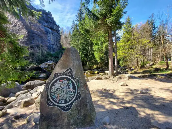 Kamień z logo Krucze Skały przy wejściu na via ferrata. W tle skalne ściany i las.